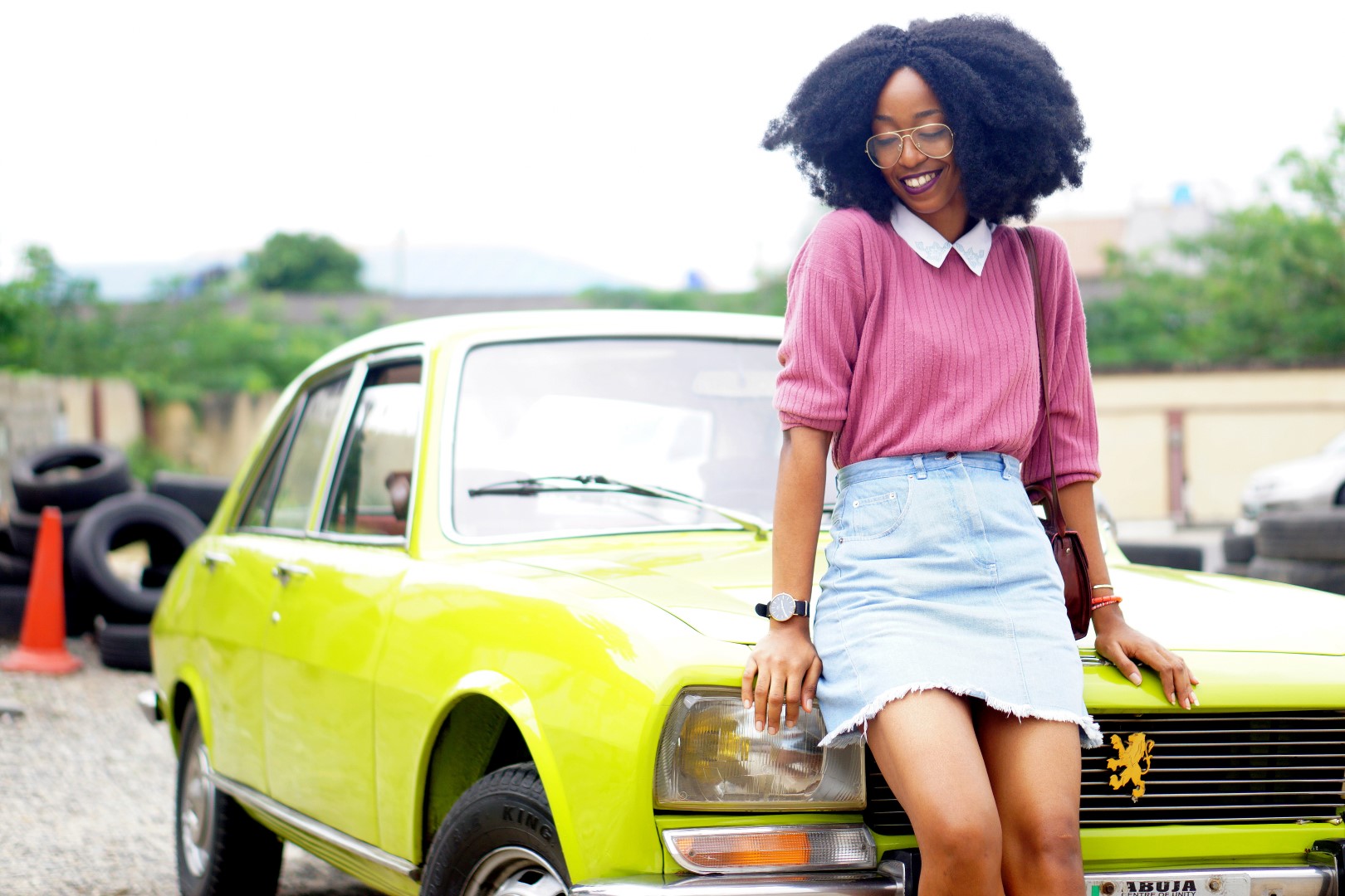 nigerian fashion blogger Cassie daves wearing a denim mini skirt, mauve sweat shirt. leaning against a lemon green peugeot 504